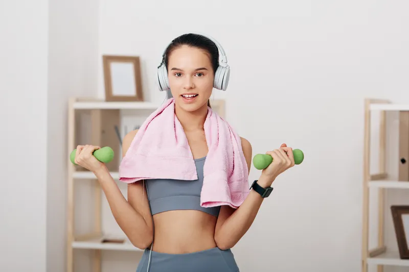 Fitness woman lifts dumbbells with headphones and a towel after a workout in a bright home gym, showcasing strength, concentration, and modern athletic lifestyle.