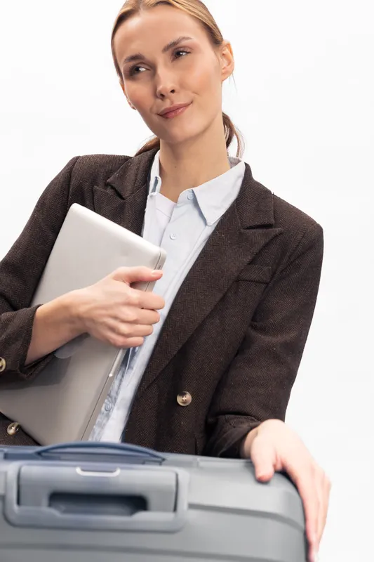 Young businesswoman in brown blazer holds laptop and leans on suitcase, looking away. Concept for travel, work, business trip, and professional lifestyle with modern technology.
