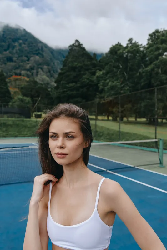 Young woman in athletic wear poses on a tennis court, showcasing confidence and grace in a vibrant outdoor setting with lush greenery and a cloudy sky