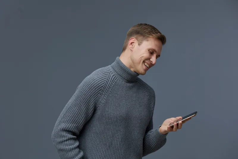 Smiling man using smartphone in casual gray sweater on plain gray background studio portrait. People lifestyle concept, technology, communication, modern, cheerful, relaxed, happy.
