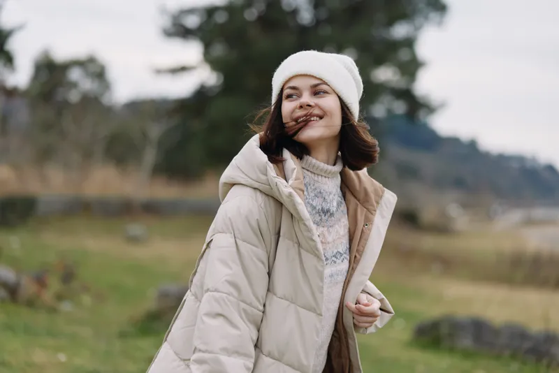 Woman in a cozy beige coat and white beanie outdoors, smiling in a wide field with trees, radiating warmth and a natural autumn vibe.