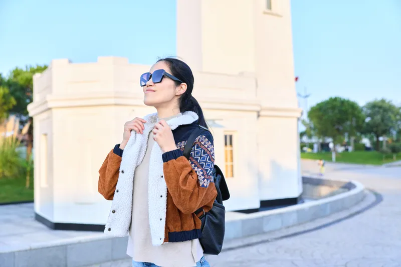 A stylish woman outdoors wearing a scarf and jacket, sunglasses and backpack, posing near a pale monument under daylight in a sunny urban setting