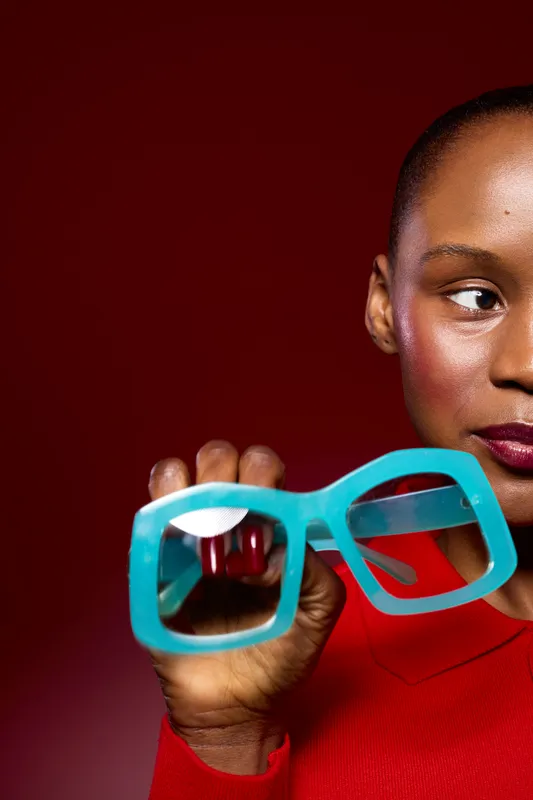 Close-up of a dark skinned African woman holding blue oversized eyeglasses against a red background. Concept of fashion, style, accessories, and modern portrait photography.