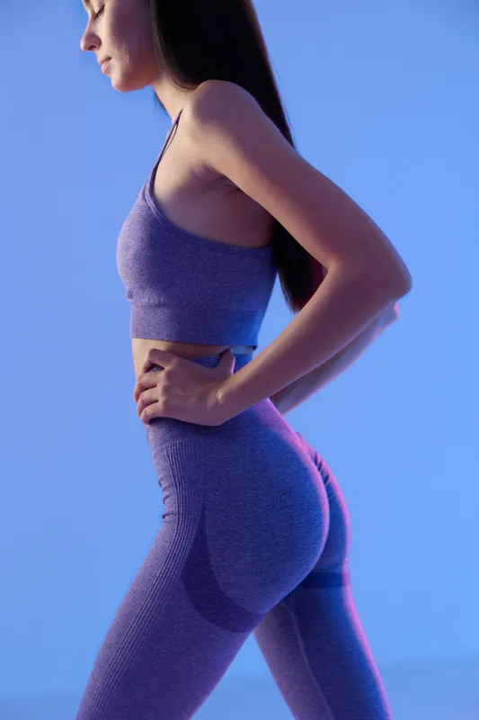 woman sport fitness in purple activewear posing in profile, slim athletic figure, long dark hair, studio lighting with blue background, focused and calm expression