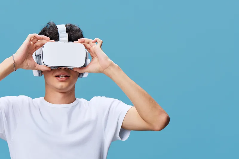 Young man using virtual reality headset with white shirt against blue background, enjoying immersive technology experience in casual style and modern setting.