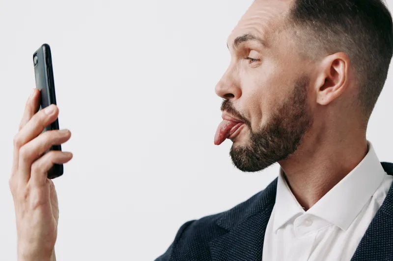 A professional man in a suit playfully sticks out his tongue while looking at a smartphone, set against an isolated colored background, conveying lighthearted emotion in a business setting.