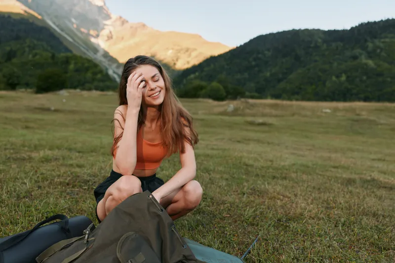 A cheerful young woman sits outdoors in a grassy meadow with distant mountains, smiling warmly while resting beside a backpack, exuding relaxed confidence and natural outdoor charm.