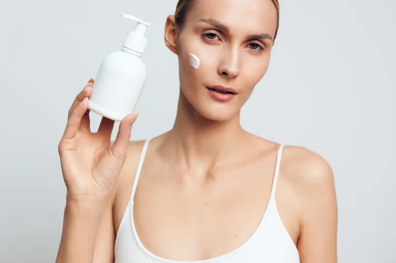 Young woman with clear skin holds white bottle with pump applies cream on face shows skincare routine on light background in white top with natural makeup and focused expression.