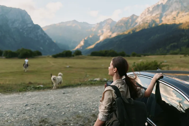 Young woman with a backpack leans on a car door in a wide mountain valley, looking toward the distant peaks and feeling ready for adventure as a gentle breeze ruffles hair and clouds drift above the