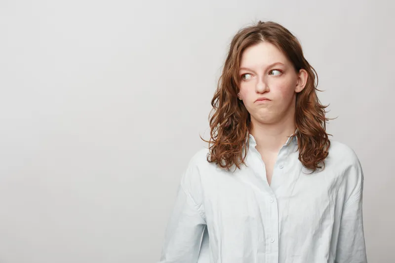 Young woman with curly hair in white shirt looks sideways with displeased expression on light background. Concept of doubt, suspicion, and negative emotions in casual portrait.