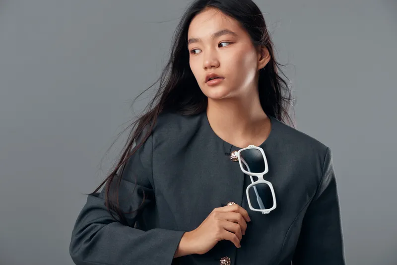 Woman, portrait and black hair holding white sunglasses with gray jacket on neutral background. Fashion, style and beauty with casual look for young female model in studio photo session.