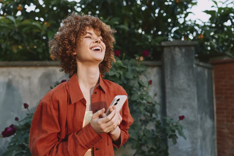 A joyful woman in an orange shirt stands outdoors in a garden, holding a smartphone with a bright smile. A lifestyle portrait capturing casual happiness