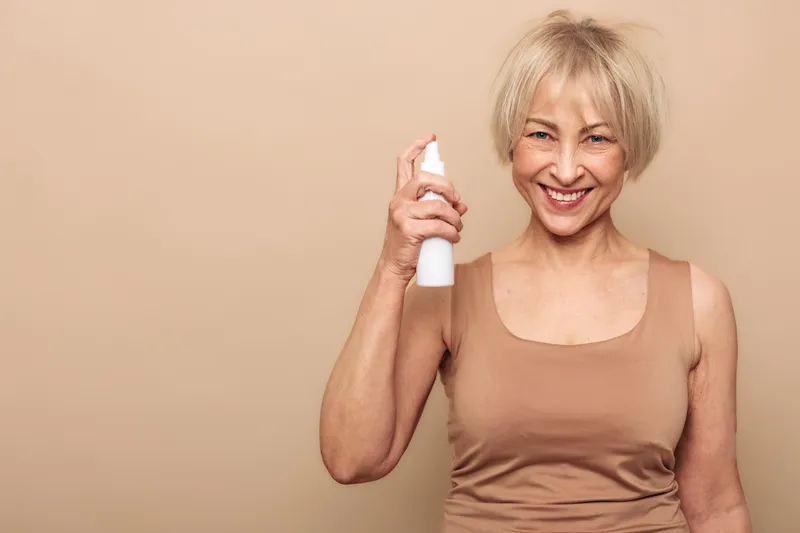 Smiling mature woman with short blonde hair holding white spray bottle, wearing beige tank top, isolated on beige background. Concept for skincare, beauty, wellness, and self-care routine.