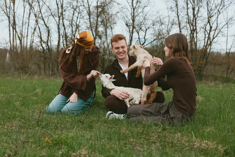 Casual group of friends outdoors with a playful goat kid, smiling and interacting in a grassy field, warm natural light, rustic setting, lifestyle moment, gentle bonding with animals, cozy and