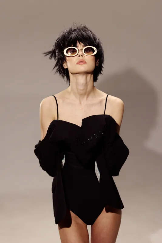 Stylish woman in black outfit wearing white sunglasses with short hair posing in minimal studio setting