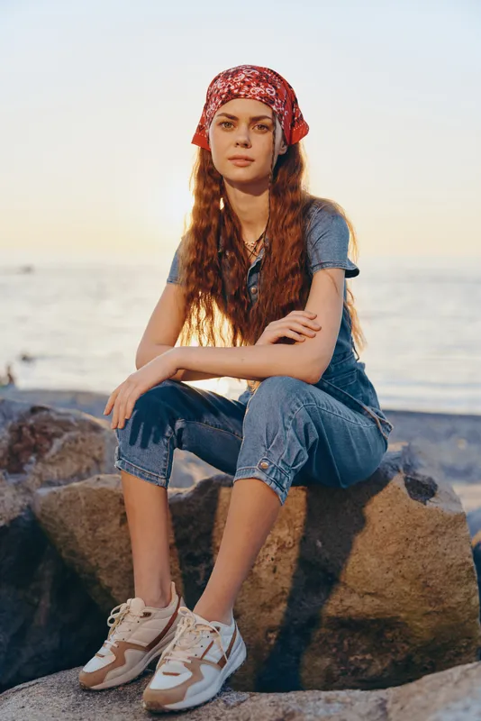Lifestyle woman in modern boho-western denim street style, wearing a red bandana and sneakers by the beach during warm sunset film color lighting.