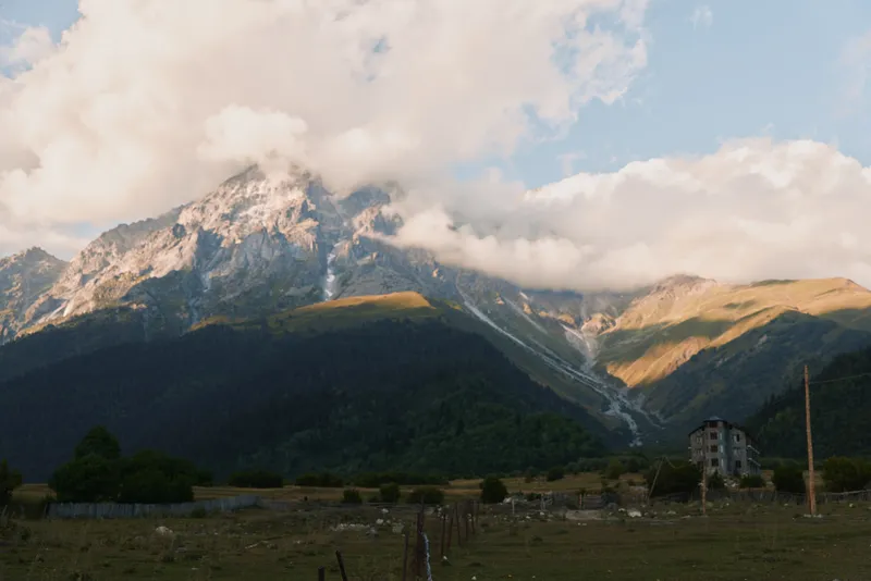 Mountain landscape with clouds covering peaks, green forest at base, grassy field in foreground, soft sunlight, peaceful rural scene, natural environment