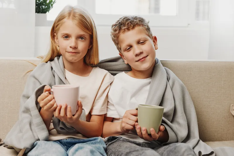 Two children wrapped in grey blankets sit on a sofa holding mugs, enjoying a cozy moment indoors. Warm natural light fills the room, conveying comfort and relaxation.