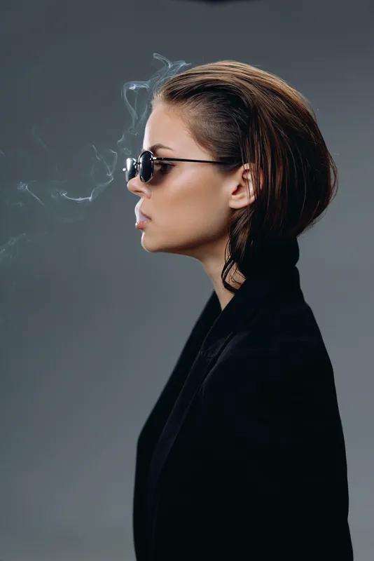Profile of a stylish woman smoking with sunglasses wearing black jacket in moody lighting and grey background