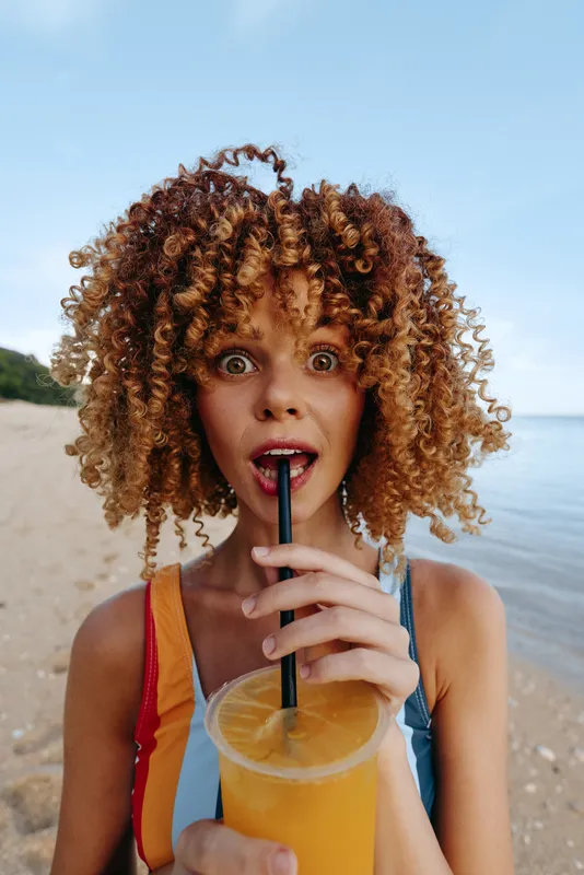 Young woman with curly red hair drinks orange juice through a straw on a sandy beach with a calm sea and blue sky in the background, showing a surprised expression.