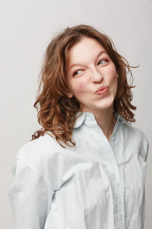 Young woman with curly hair wearing white shirt makes playful face looking sideways. Studio portrait on light background. Concept of fun, youth, and casual lifestyle.