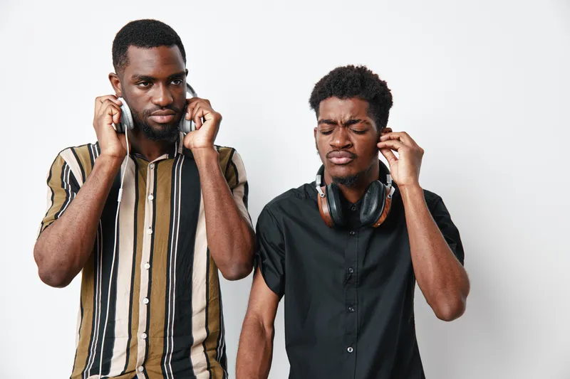 Two african men with dark skin wearing headphones standing against white background. One man in striped shirt and other in black shirt with eyes closed and focused expression.