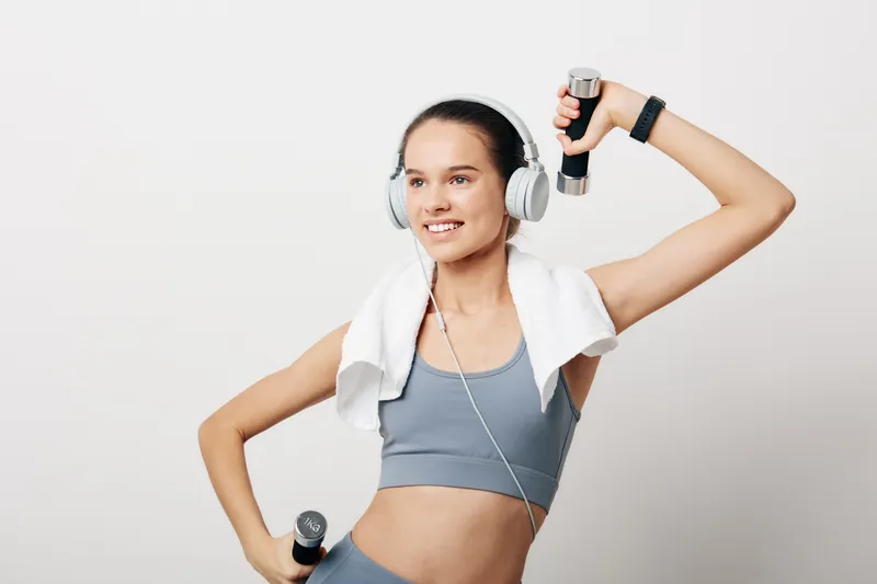 Fitness woman in sport attire lifts dumbbells with a towel over her shoulders and headphones, mid workout in a bright studio, radiating energy and focused training.