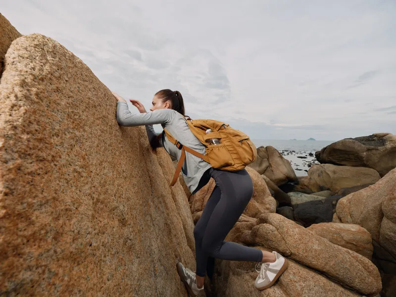 Young woman climbing rocky terrain by the sea, showcasing adventure with a backpack, engaged and focused in a natural outdoor setting