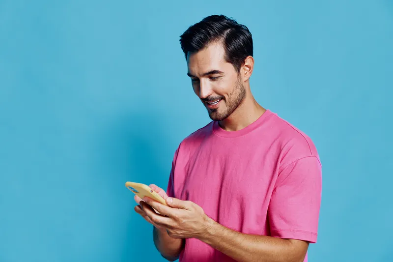 Smiling man wearing pink t-shirt looking at smartphone screen, enjoying social media, isolated on solid blue background. Positive lifestyle concept.