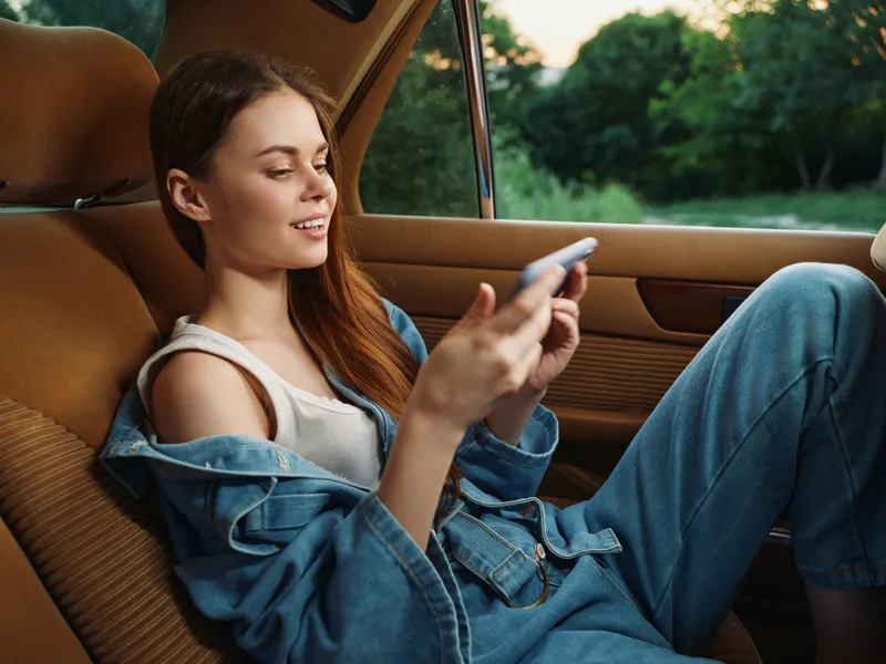 Young woman relaxing in a car, smiling and using a smartphone, wearing a denim jacket and jeans in a natural outdoor setting at sunset.