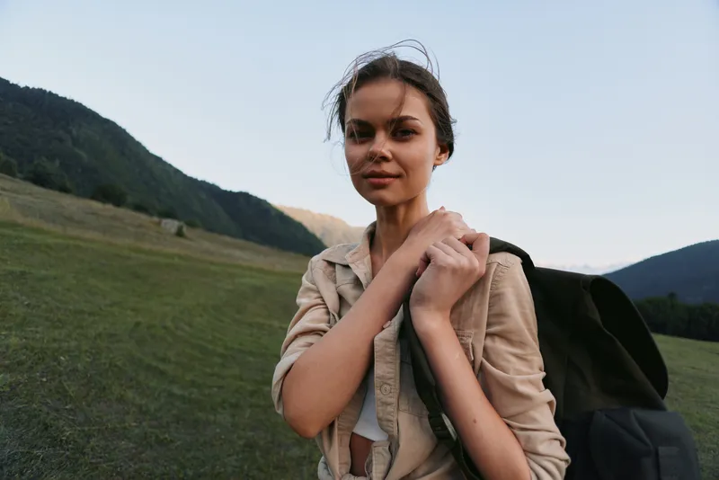 Woman traveler outdoors with backpack in a meadow, mountains visible, casual hike mood, natural light, serene landscape, lifestyle portrait in nature and exploration