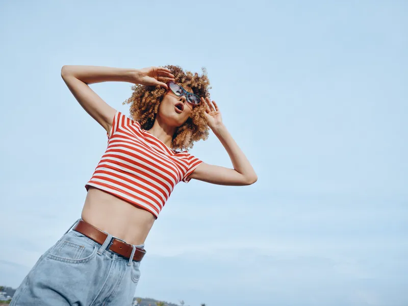 A stylish young woman poses outdoors under a clear blue sky, wearing a red and white striped crop top and high waisted jeans, exuding youthful confidence and effortless fashion.