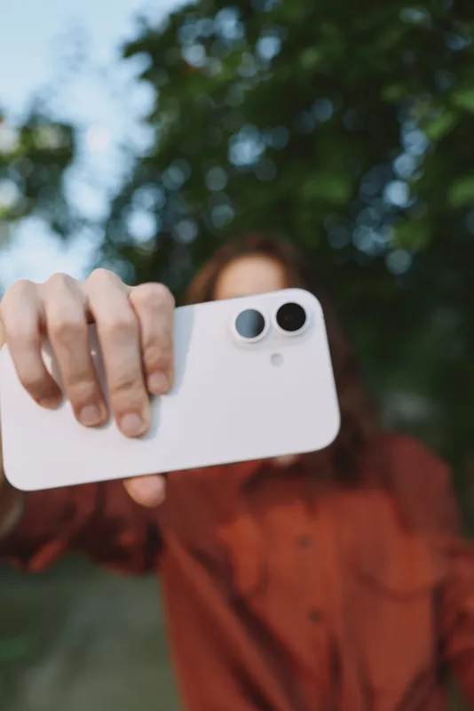 Young woman takes a casual outdoor selfie with a smartphone, wearing a rust shirt, natural light, green park background, candid lifestyle portrait, relaxed mood, in frame