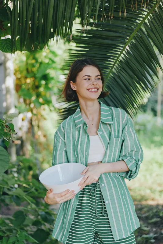 Smiling woman outdoors under palm leaves, holding a white bowl, wearing a green striped shirt and light jacket in a sunny garden.