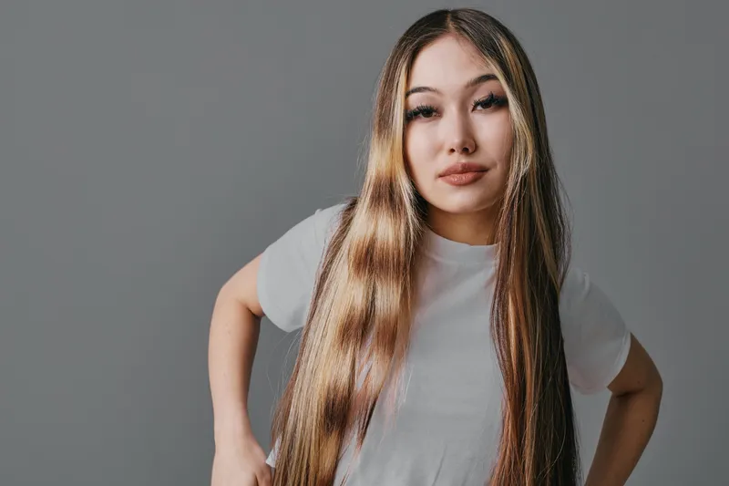 Young woman with long straight hair and neutral expression stands against grey background wearing white t shirt casual style and natural makeup indoors.
