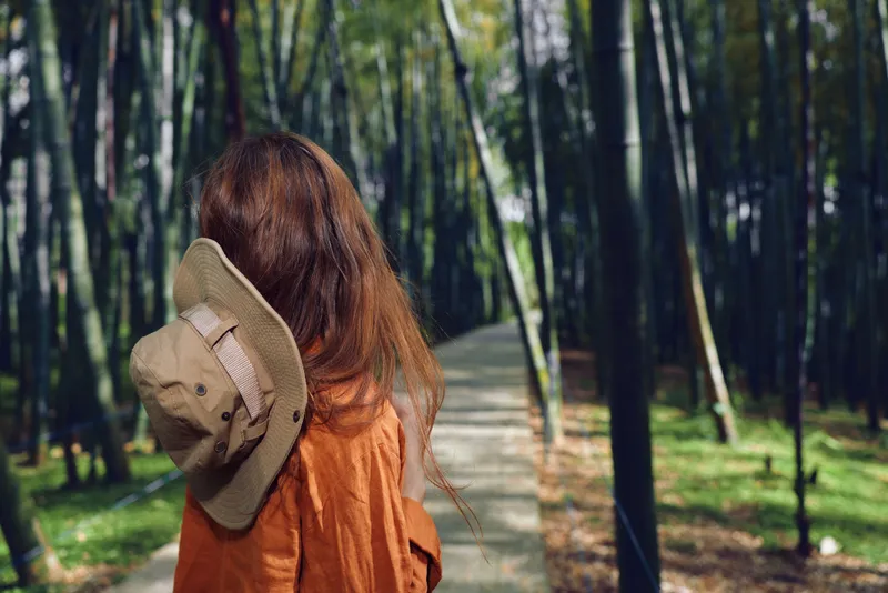 Woman with hat and long hair seen from back walking along a paved path through dense bamboo forest, wearing rust orange coat, travel mood, nature trail, serene peaceful outdoor scene for wander