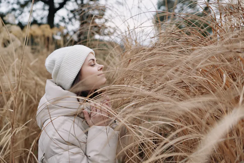 woman, winter, nature, grass, outdoor, coat, serenity, portrait, A woman in a white coat and knit cap leans toward soft beige grasses, embracing quiet outdoor solitude and a gentle, cold breeze in a
