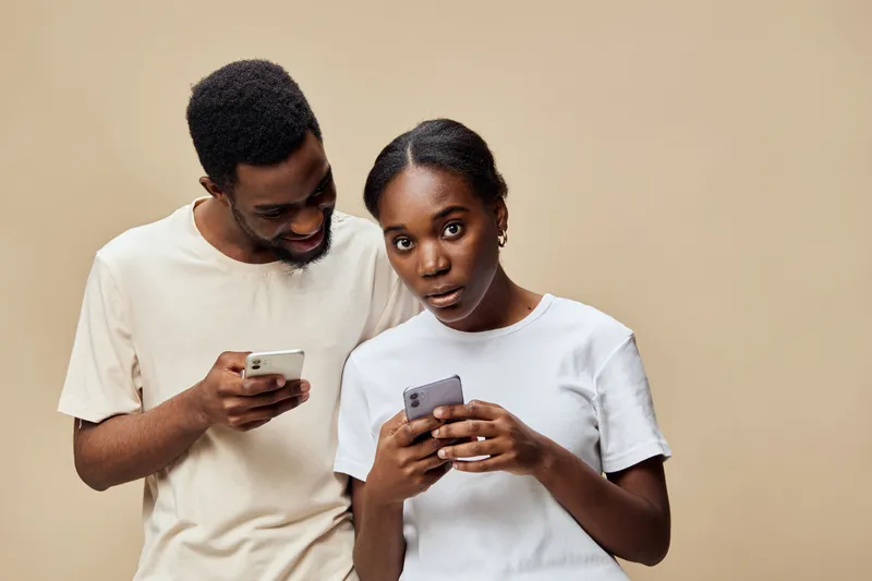 Young African couple engaging with smartphones on a neutral background, showcasing modern technology use, curiosity, and connection
