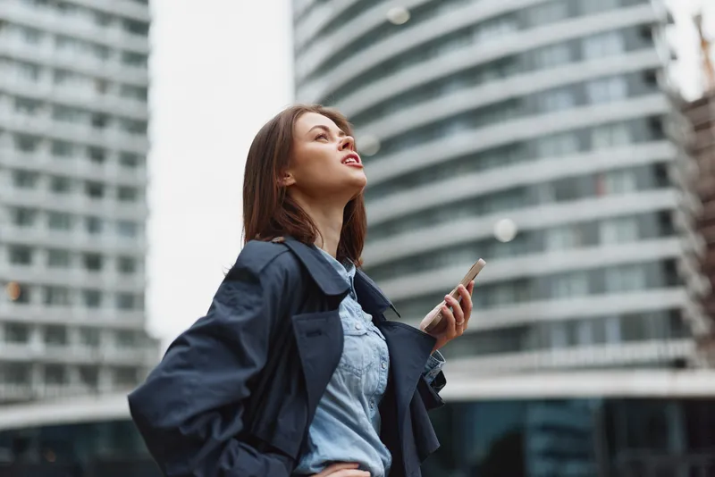 Young woman with long brown hair stands confidently outdoors in a blue jacket holding a smartphone while looking up with a thoughtful expression.