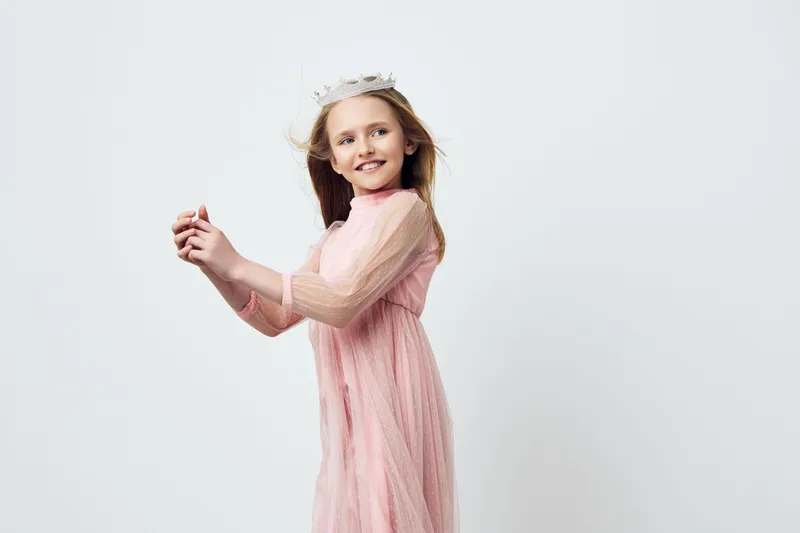 Cute girl in a pink dress with a crown, smiling and playing joyfully against a neutral background, perfect for children s fashion and celebration concepts