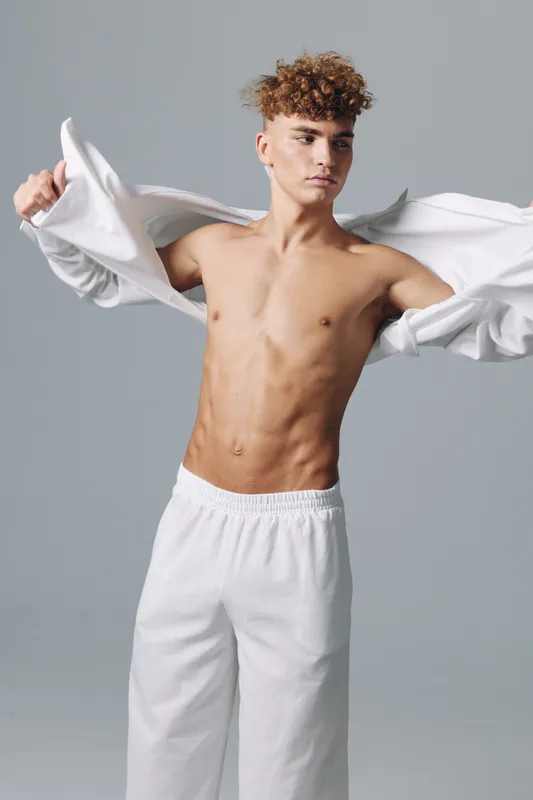 Young man with curly hair taking off white shirt showing toned bare chest and wearing white pants in studio with neutral background. Fashion, style and casual clothing concept.