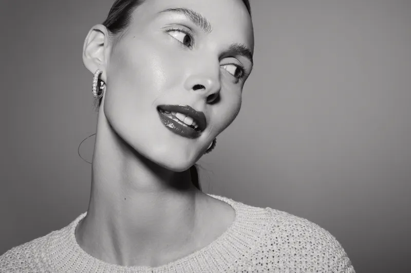 Portrait of a young white woman with fair skin, wearing earrings and a knitted sweater, looking sideways with glossy lips in monochrome studio light.