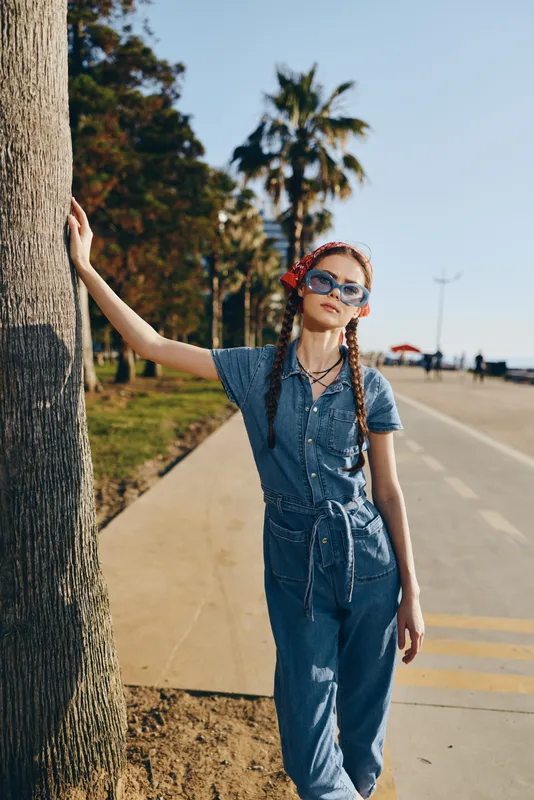 Lifestyle woman in modern boho-western denim street style with sunglasses and braided hair posing confidently near palm trees under film color tones.