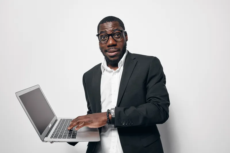 Black man with dark skin wearing glasses and black suit holding laptop and typing on keyboard isolated on white background for technology and business concept.