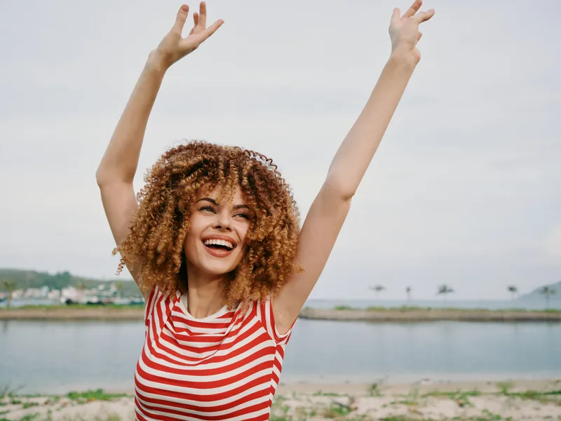 Joyful woman with curly hair wearing a striped shirt raises arms in celebration on a beach, capturing a bright, candid moment of freedom and radiant expression.