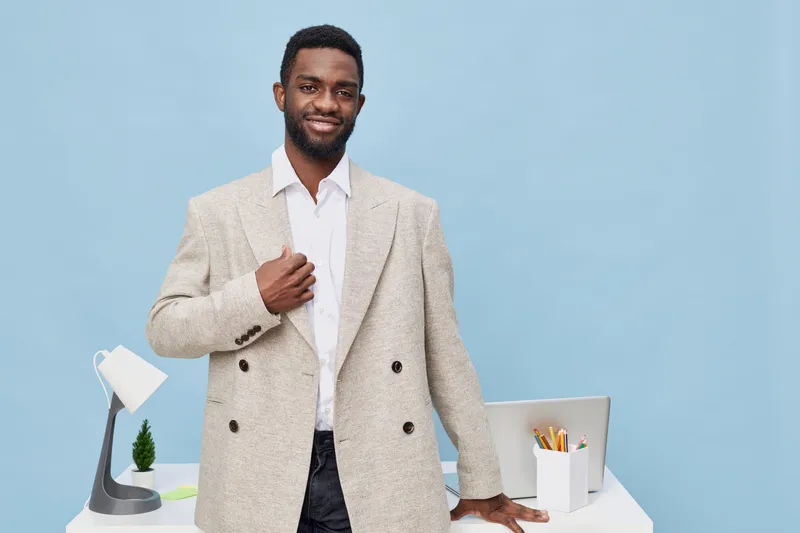 Confident smiling man in beige blazer and white shirt standing at modern white office desk with lamp, pencil holder and computer, professional businessman in creative workspace, positive mood. People 