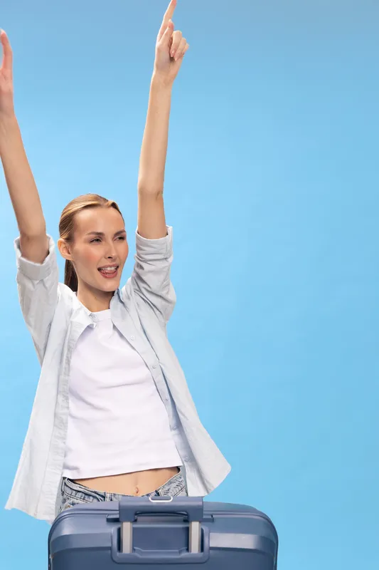 Young happy woman in casual clothes raises hands up excitedly behind suitcase. Blue background studio portrait of traveler celebrating vacation or trip with joyful expression and positive mood.