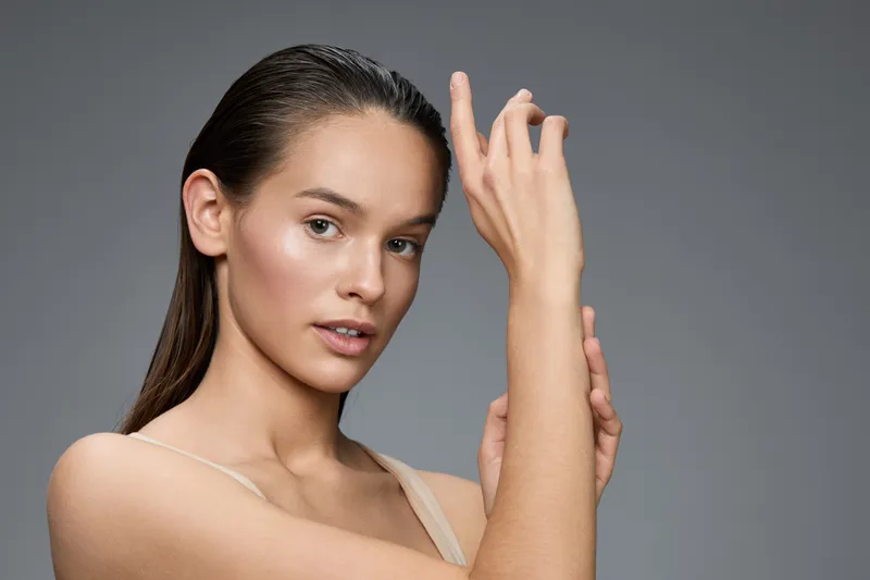 Young woman with natural makeup and wet hair poses gracefully against a neutral gray background, showcasing elegance and confidence through beauty and self care