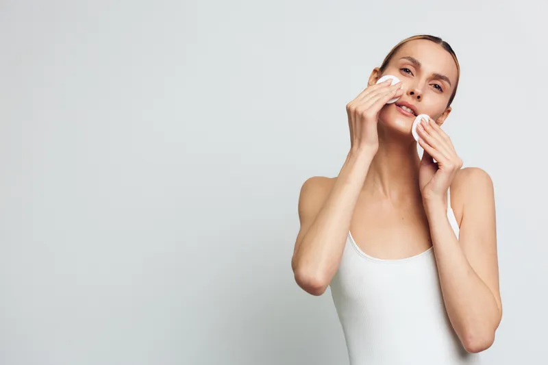 Young woman with clean skin removing makeup with cotton pads on a light background. Concept of skincare, beauty routine, and facial cleansing. Soft natural lighting highlights fresh skin.