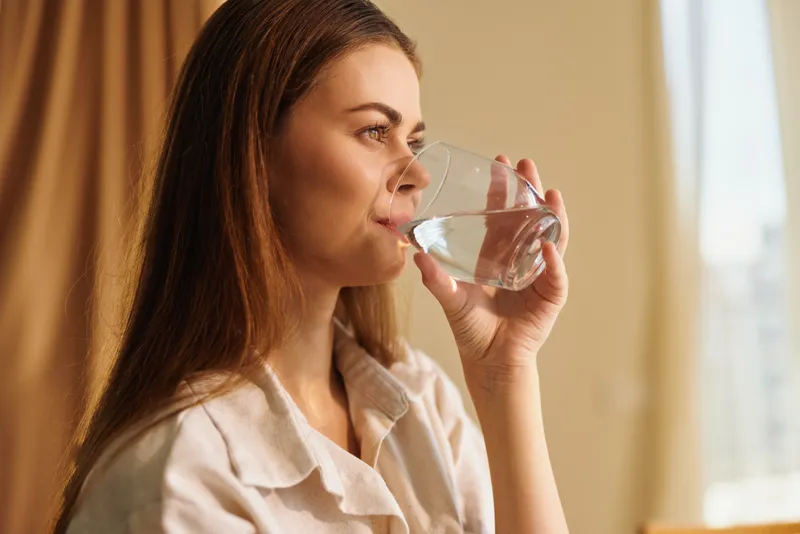 Woman drinking water from a glass at home in the morning, portrait closeup showing a healthy routine, natural light through window, casual shirt, relaxed and thoughtful expression.